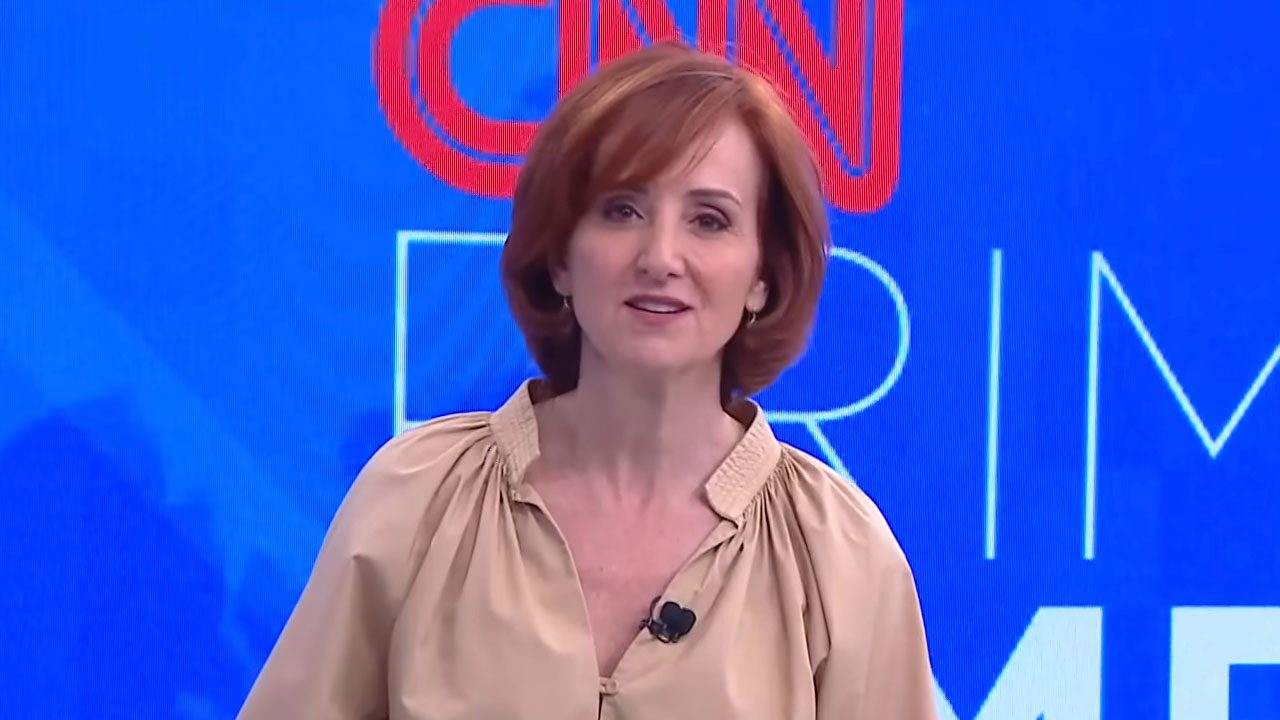 Thais Herédia no estúdio da CNN Brasil, vestindo uma camisa bege, com o cabelo ruivo e fundo azul do programa