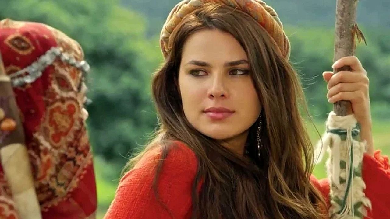 Mulher com traje típico e lenço na cabeça segura cajado em cenário rural com expressão desconfiada