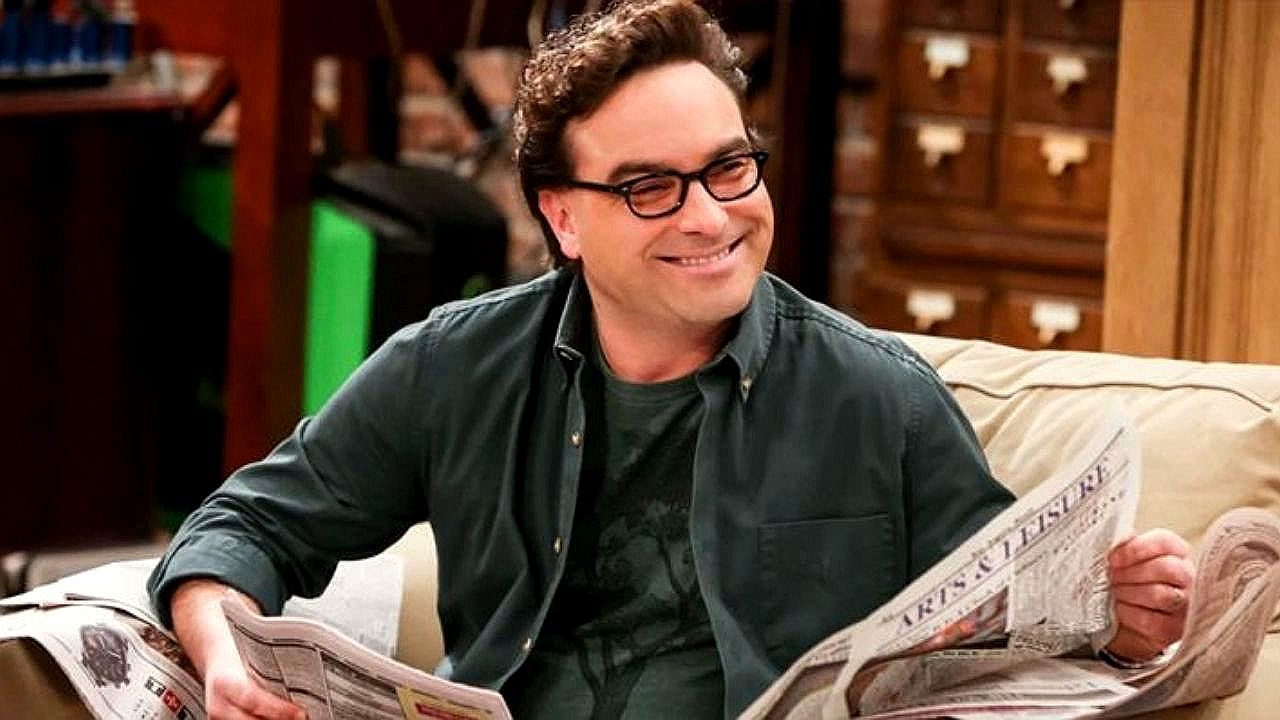Leonard Hofstadter sorri no sofá enquanto lê jornal em cena da série The Big Bang Theory