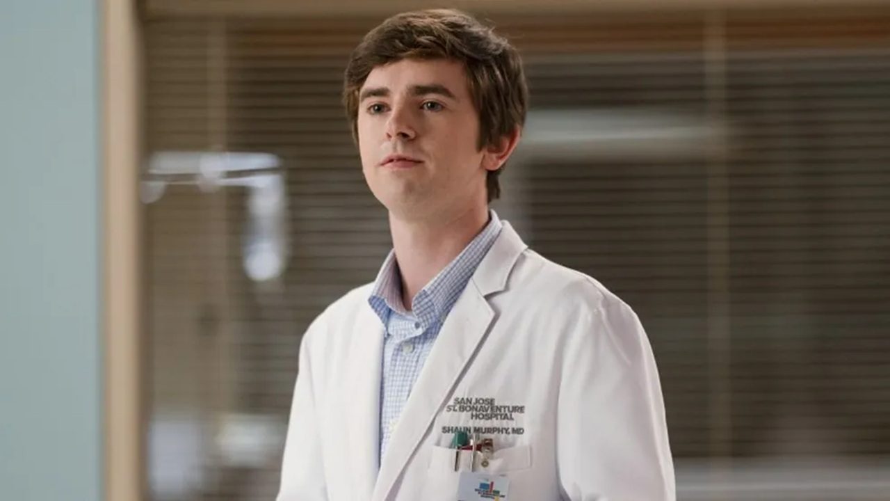 Homem jovem veste jaleco branco com crachá de hospital e expressão atenta, em ambiente clínico com paredes claras e janelas ao fundo