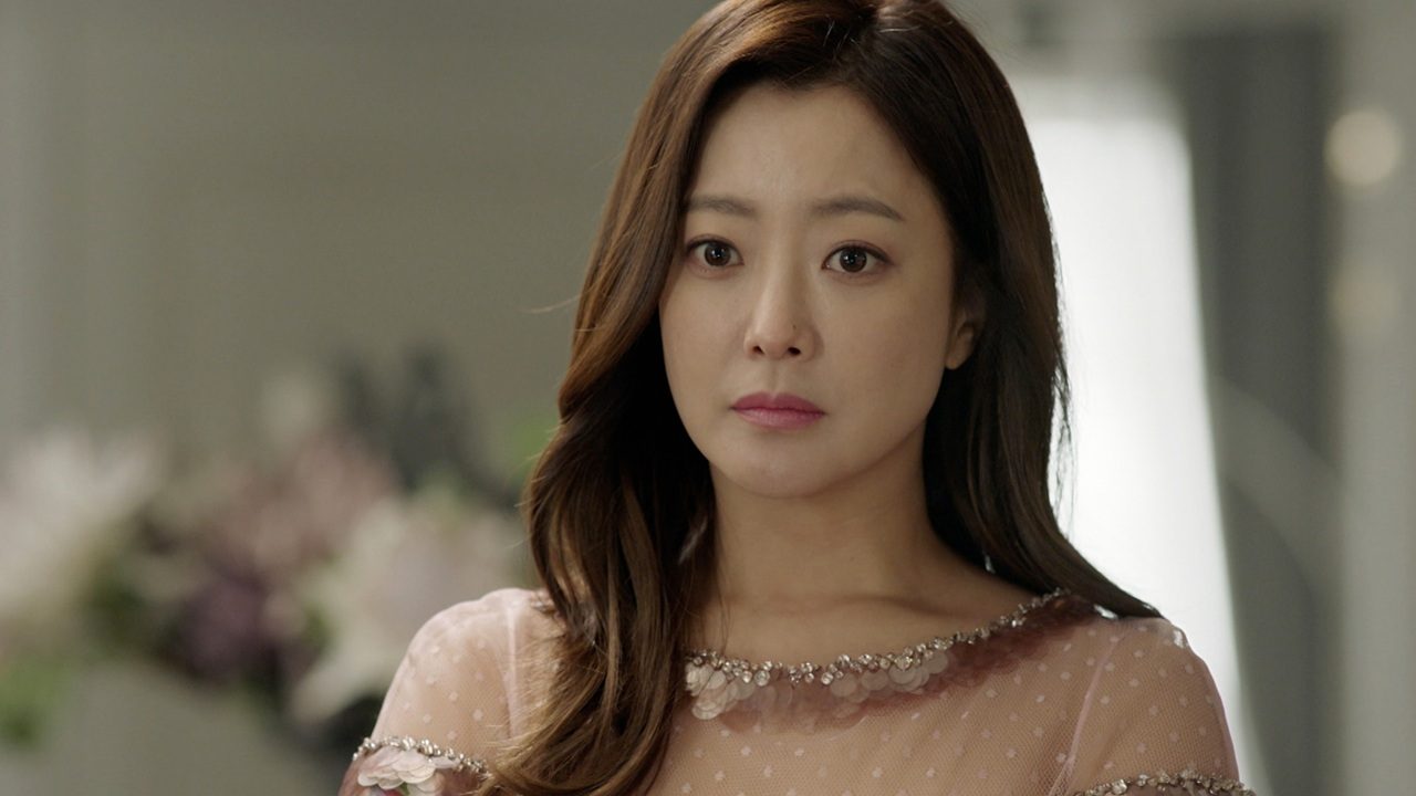 Mulher com expressão séria olha à frente em ambiente interno iluminado; ela tem cabelo castanho ondulado e veste um vestido claro com detalhes bordados e transparências sutis.