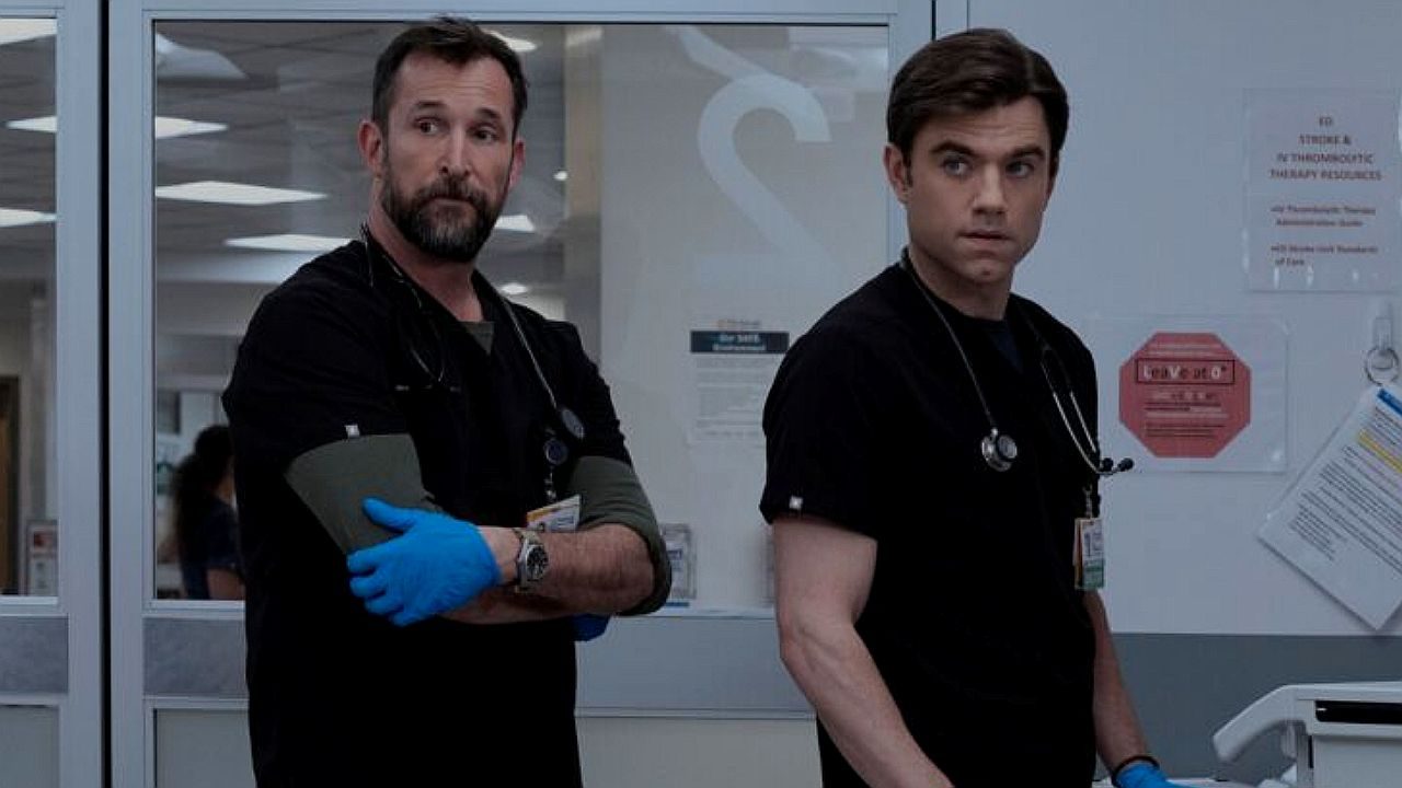 Dois profissionais de saúde vestindo uniformes médicos pretos e com estetoscópios no pescoço estão em um corredor de hospital. Um deles usa luvas azuis e mantém os braços cruzados, enquanto ambos olham para o lado com expressão séria, diante de portas de vidro e avisos fixados na parede, sugerindo momento de tensão ou atenção a uma situação clínica.