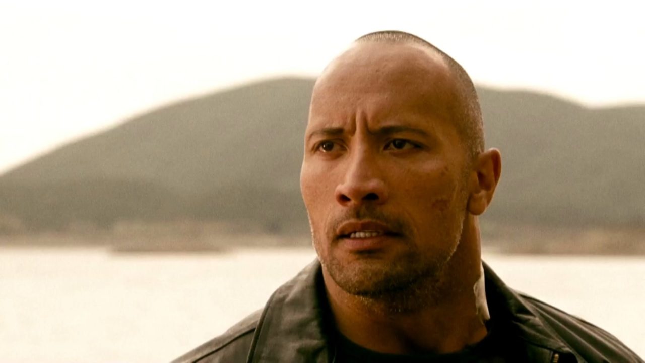 Driver (Dwayne Johnson) com expressão séria e jaqueta escura diante de lago e montanhas em cena de Rápida Vingança, filme exibido pela Band