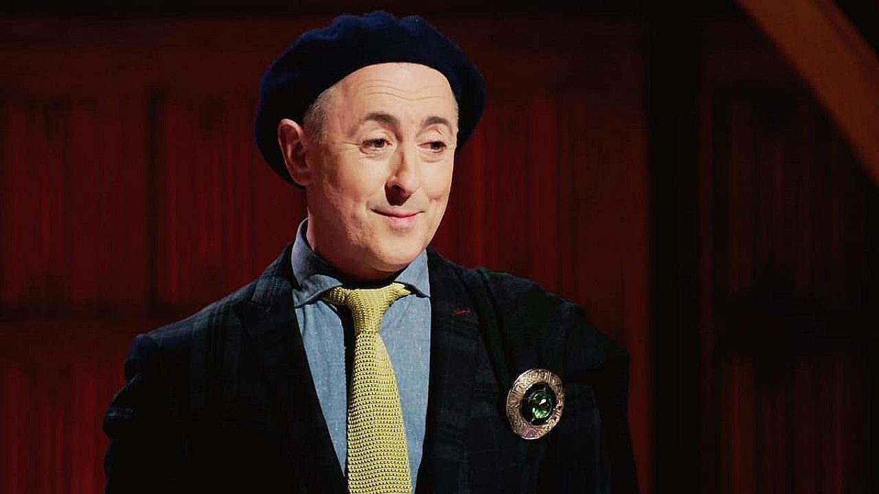 Homem usa boina preta, camisa azul e gravata amarela, combinadas com blazer escuro que traz um broche circular na lapela. Ele aparece em ambiente interno com fundo de madeira, exibindo expressão leve e discreto sorriso, como em cena de programa ou apresentação.