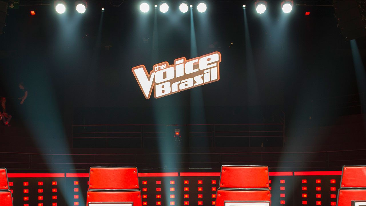 Cadeiras vermelhas iluminadas no palco com logotipo do The Voice Brasil ao fundo