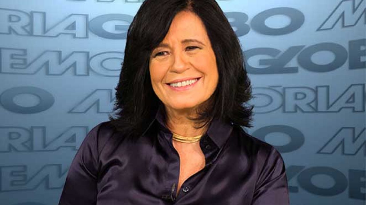 Thelma Guedes sorrindo em entrevista nos estúdios da Globo, com fundo azul escrito "Memória Globo".