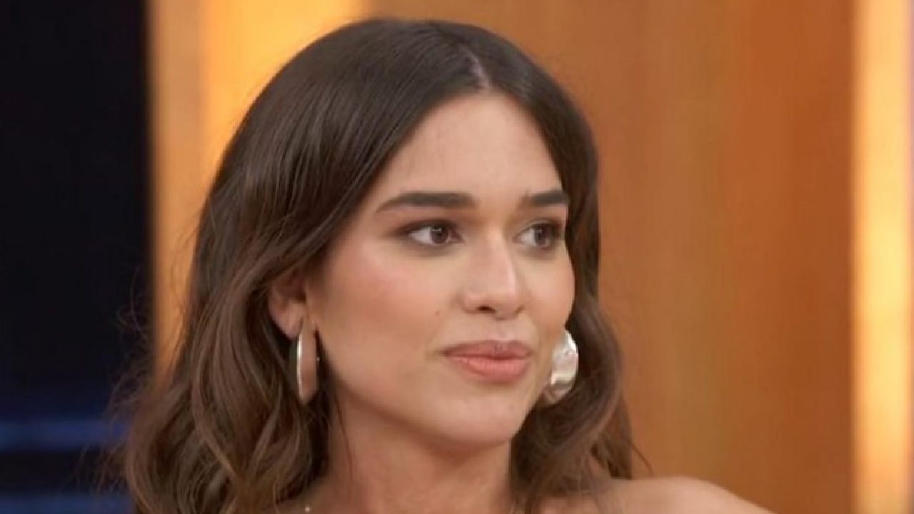 Mulher com cabelos castanhos ondulados e brincos grandes posa em estúdio de TV, com expressão atenta e fundo iluminado em tons quentes.