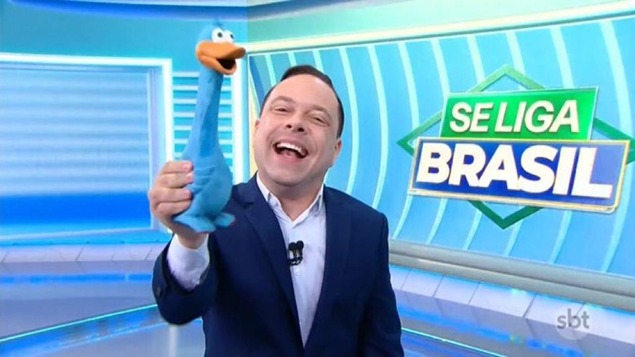 Thiago Gardinali, apresentador do Se Liga, Brasil, sorri no estúdio do SBT enquanto segura fantoche azul diante do telão do jornalístico.