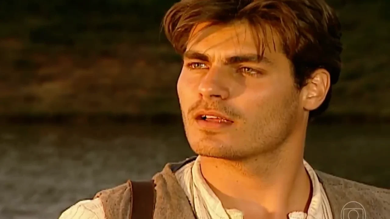 Homem jovem com cabelos castanhos e barba rala aparece em close sob a luz dourada do pôr do sol. Ele olha para o horizonte com expressão séria. Veste camisa clara de linho e colete rústico, com uma alça de mochila sobre o ombro. Ao fundo, vê-se um rio desfocado e vegetação escura