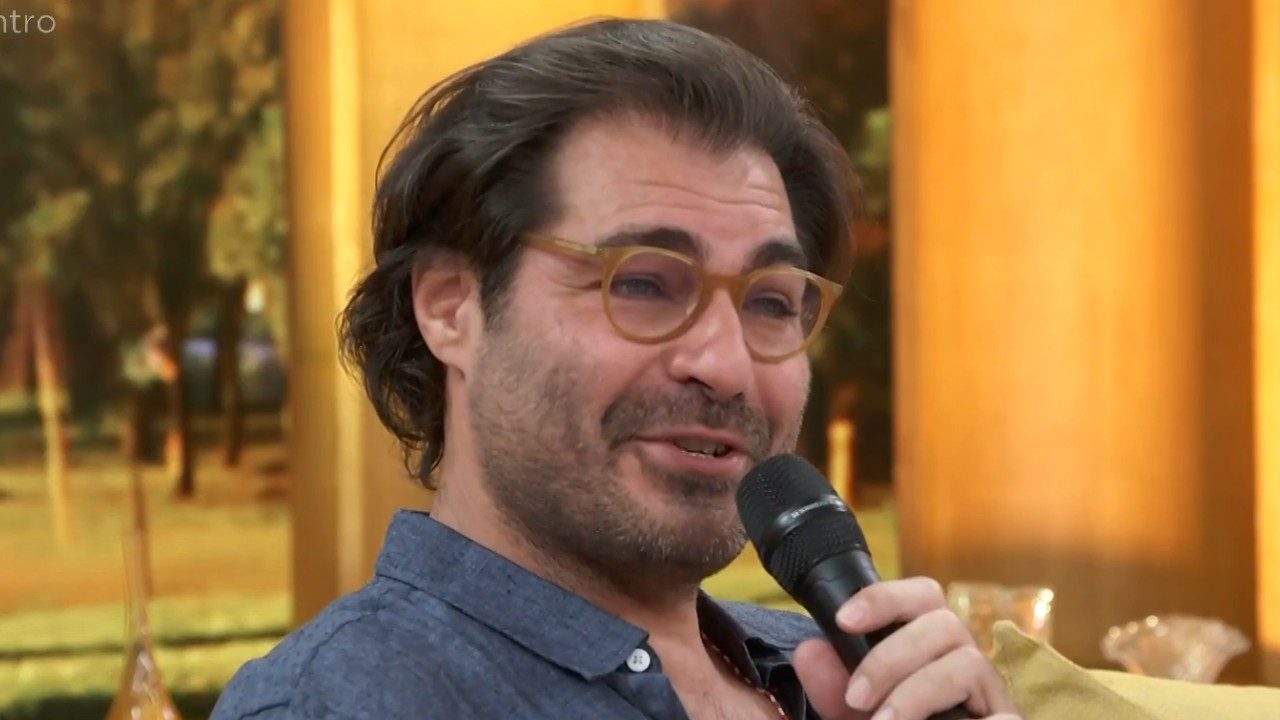 Thiago Lacerda fala ao microfone durante participação no programa Encontro com cenário iluminado ao fundo