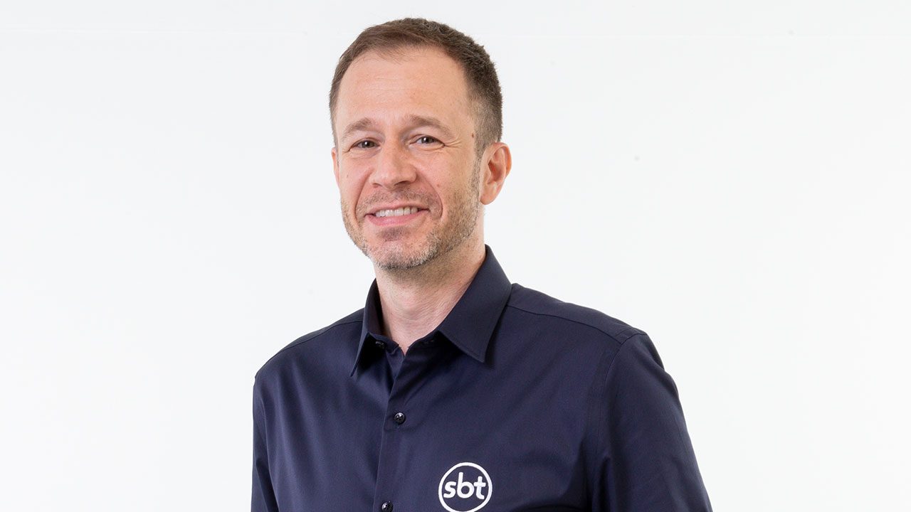 Tiago Leifert sorrindo com camisa azul marinho do SBT em foto de divulgação com fundo branco