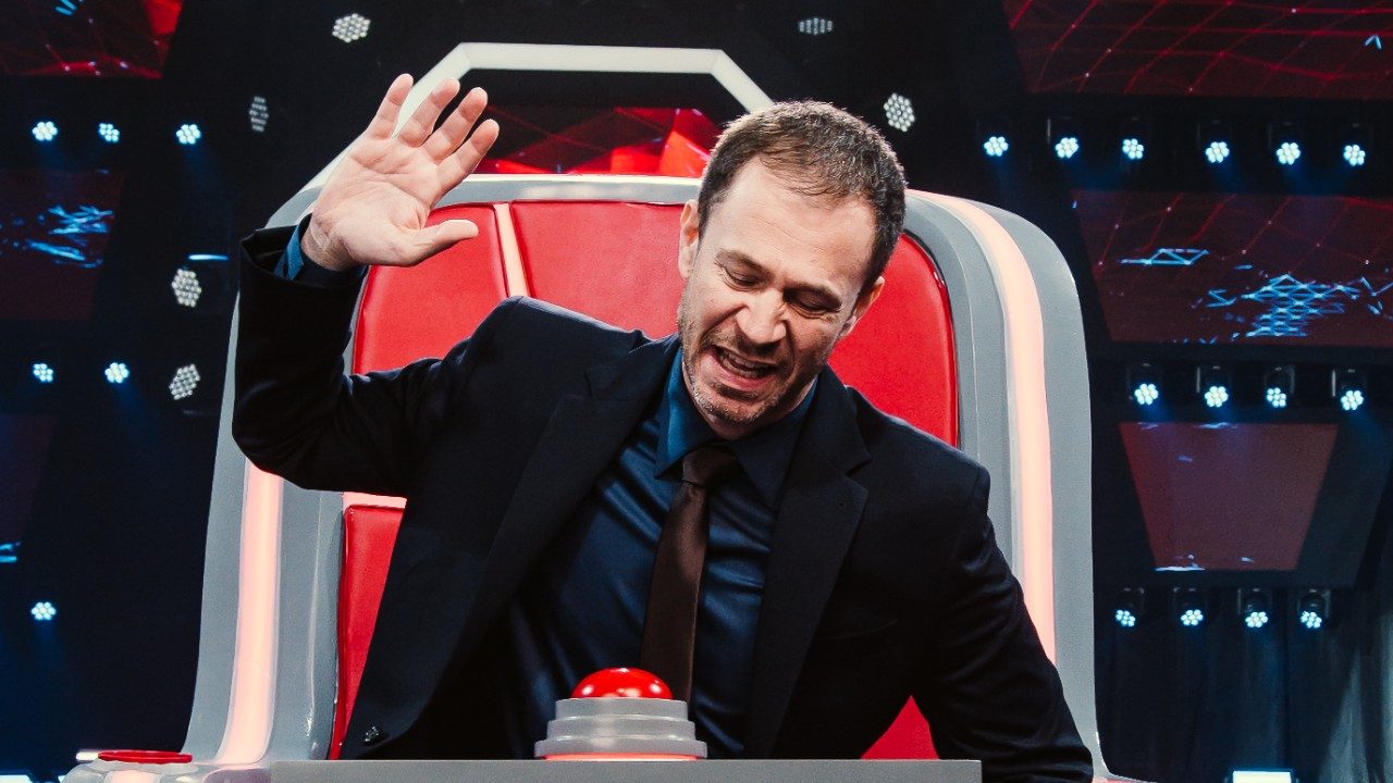 Tiago Leifert aperta botão na cadeira do The Voice Brasil com mão erguida em estúdio