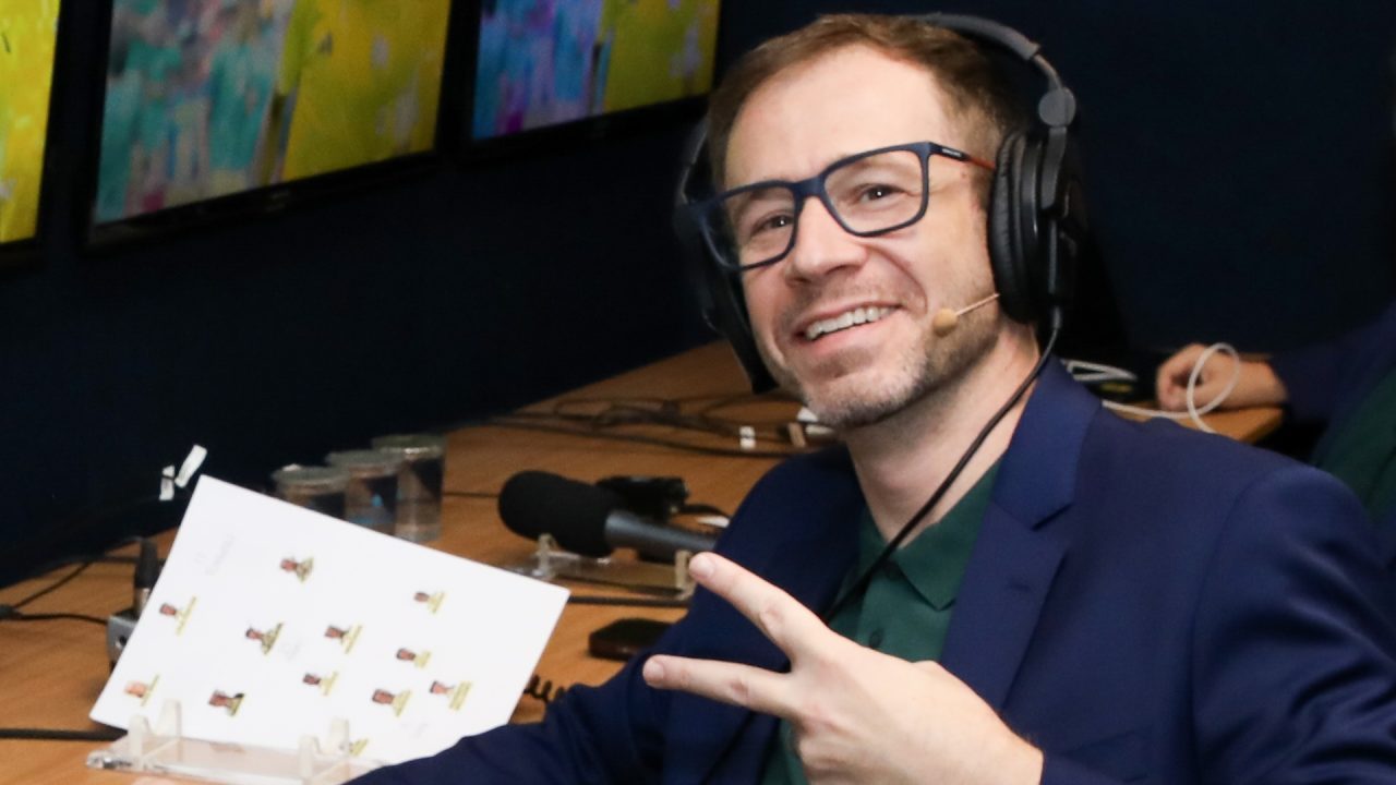 Tiago Leifert, de blazer azul e camisa verde, sorri na cabine de narração do SBT com headset e microfone durante México x África do Sul