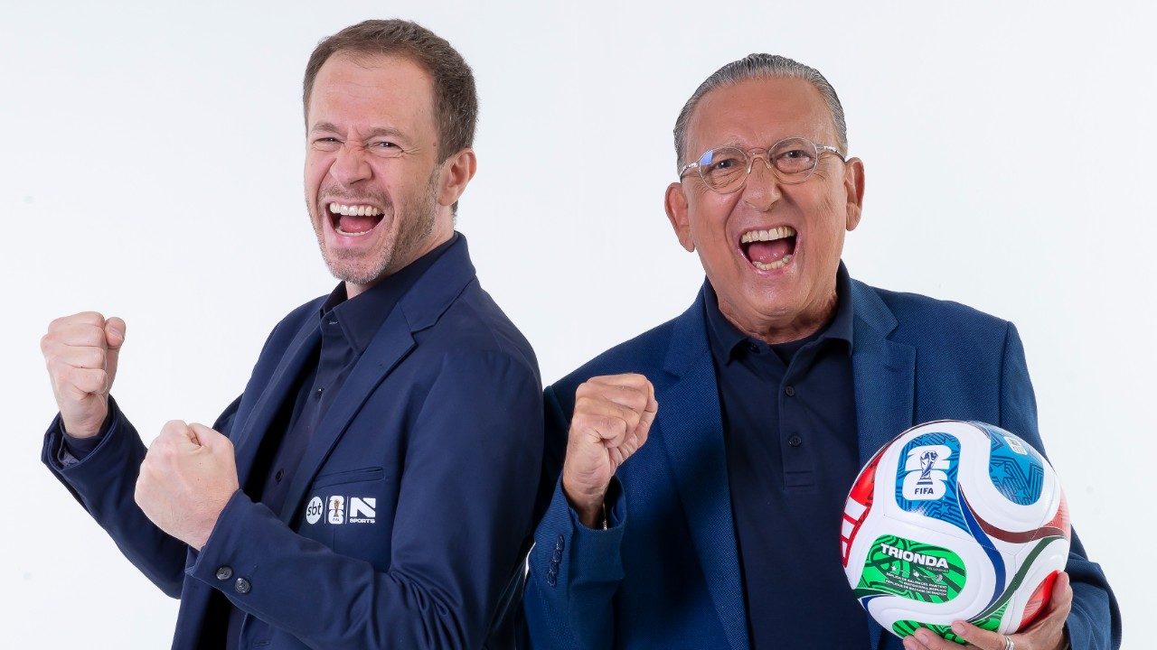 Tiago Leifert e Galvão Bueno de blazer azul comemoram com punhos cerrados enquanto posam com bola de futebol em foto promocional do SBT