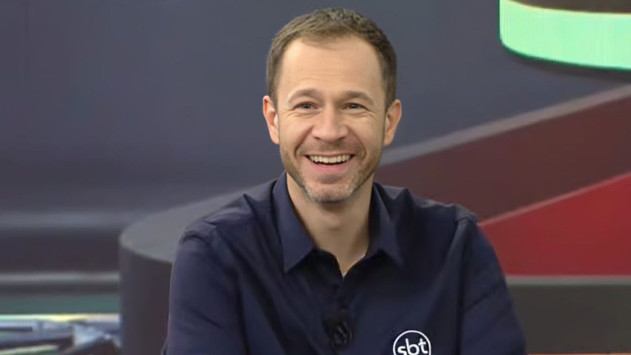 Tiago Leifert, apresentador do SBT, sorri durante participação em programa de TV em estúdio com cenário colorido ao fundo.