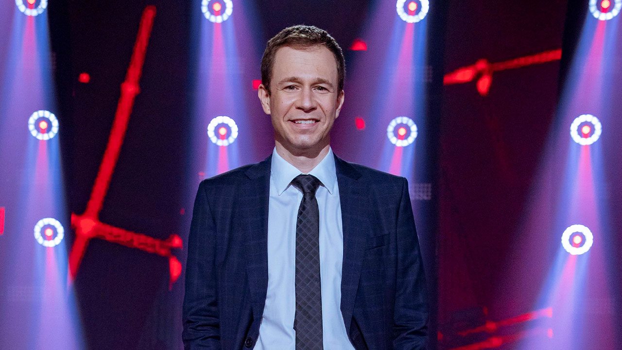 Tiago Leifert de terno escuro e gravata preta no palco com iluminação de luzes vermelhas ao fundo