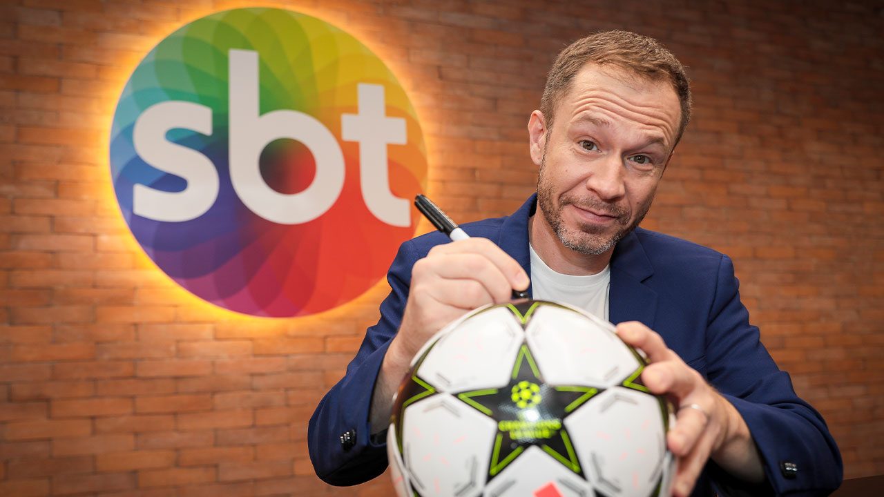 Tiago Leifert de blazer azul autografando uma bola de futebol com caneta preta, em frente ao logotipo colorido do SBT