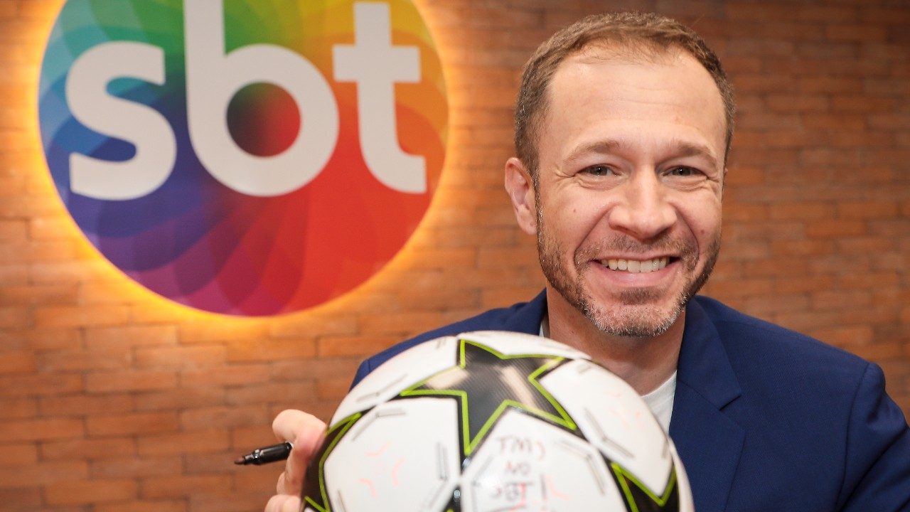 Tiago Leifert sorri com bola de futebol autografada em frente ao logotipo do SBT