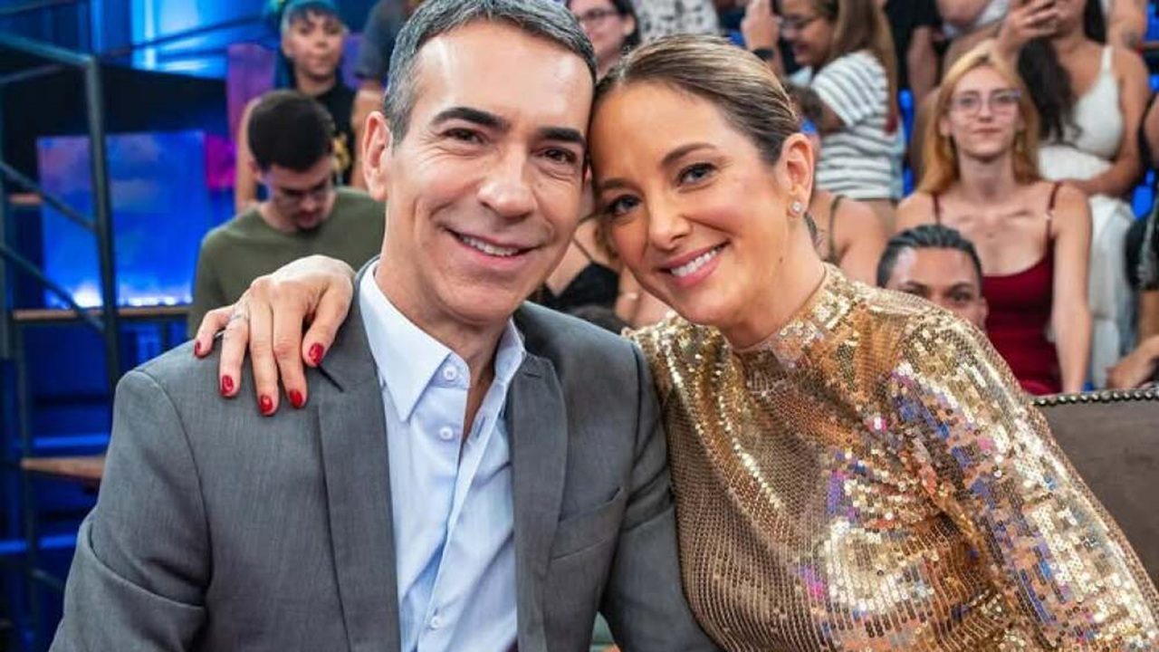 Casal posa sorrindo em um estúdio de TV, sentado diante de uma plateia. A mulher, usando um vestido dourado de paetês, abraça o homem, que veste terno cinza. Ambos estão próximos e parecem felizes, enquanto o público ao fundo observa e tira fotos