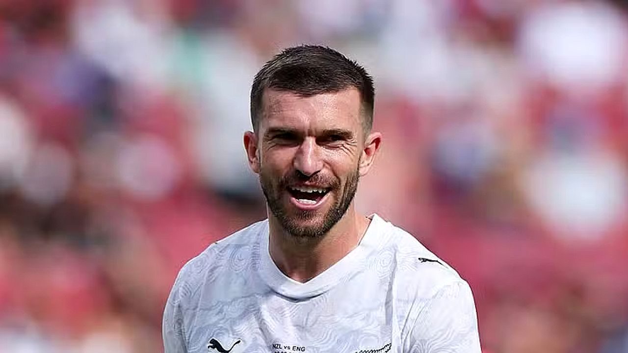 Jogador de futebol de uniforme branco sorri durante partida em estádio com arquibancadas desfocadas ao fundo e expressão de comemoração