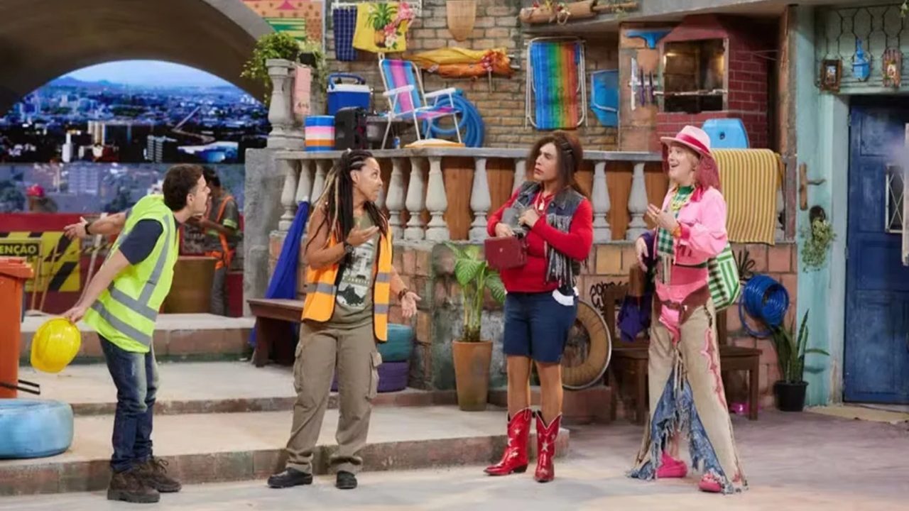 Quatro pessoas estão em um cenário colorido que simula uma vila urbana, com casas simples, varandas, plantas, objetos domésticos e vista de uma cidade ao fundo. À esquerda, um homem jovem de pele clara usa colete refletivo amarelo sobre camiseta verde, calça jeans e botas, segura um capacete amarelo na mão e inclina o corpo para frente em atitude curiosa enquanto fala com os outros. Ao centro esquerdo, uma mulher negra de estatura baixa usa colete laranja de segurança sobre camiseta escura, calça cargo bege e botas, tem cabelo longo em tranças e gesticula com as mãos enquanto conversa, demonstrando surpresa ou explicação.