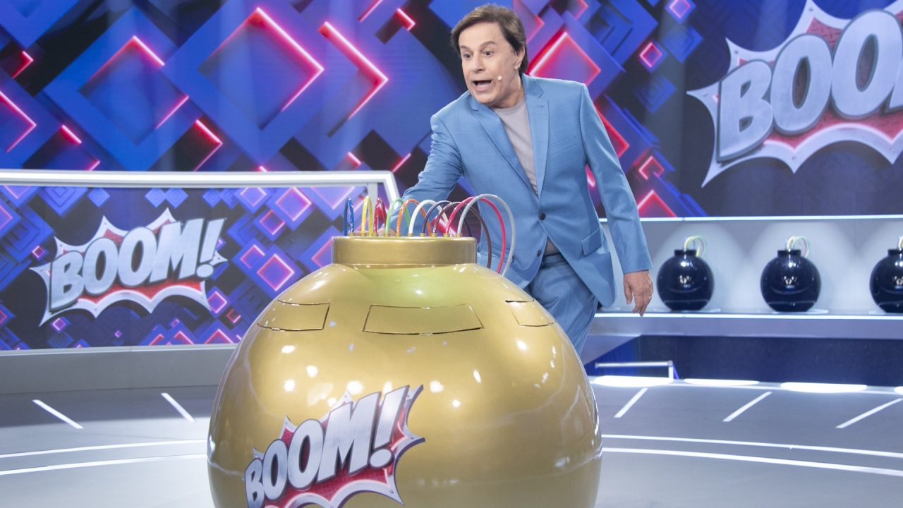 Tom Cavalcante, vestindo um terno azul, interage com um grande dispositivo dourado com fios coloridos em um estúdio de programa de auditório. Ao fundo, painéis luminosos exibem a palavra Boom! e prateleiras com objetos em formato de bomba decoram o cenário.