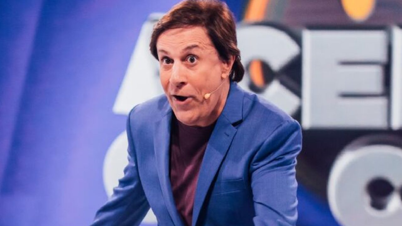 Tom Cavalcante com expressão divertida usa blazer azul e microfone de lapela, em cenário de programa de televisão com fundo iluminado e logotipo desfocado
