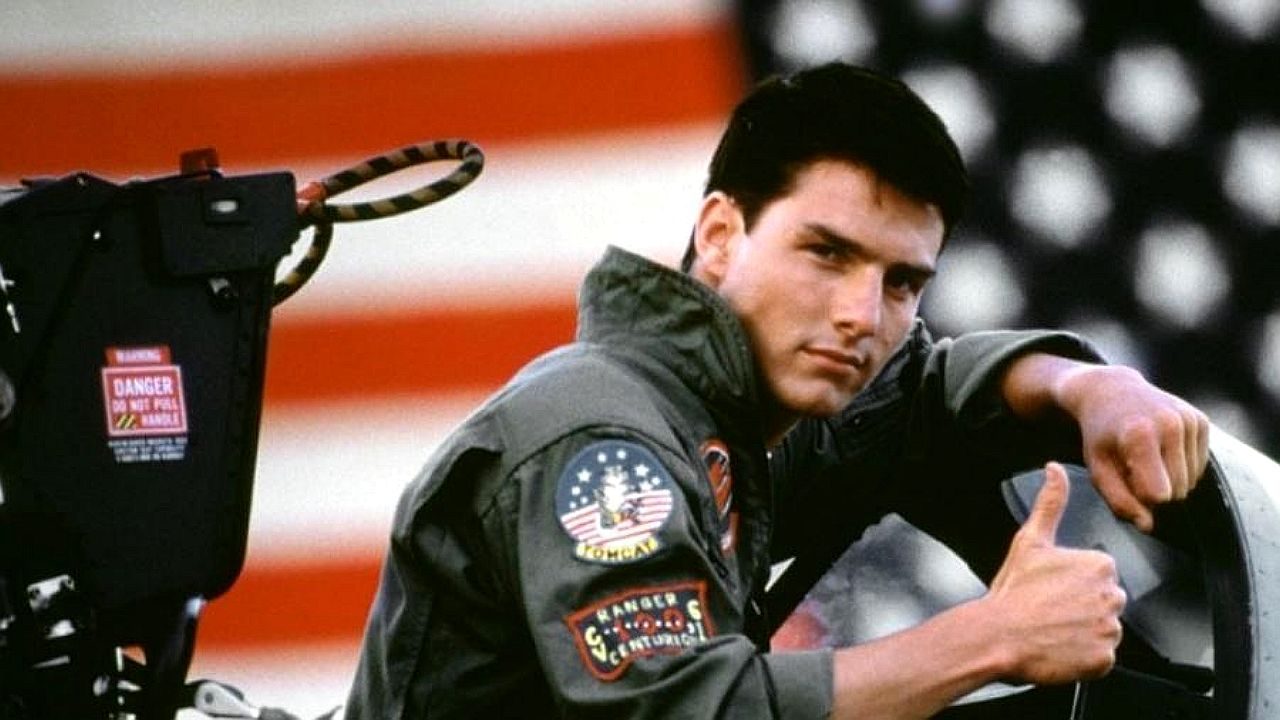 Piloto com uniforme militar faz sinal positivo ao lado de avião em cena do filme Top Gun