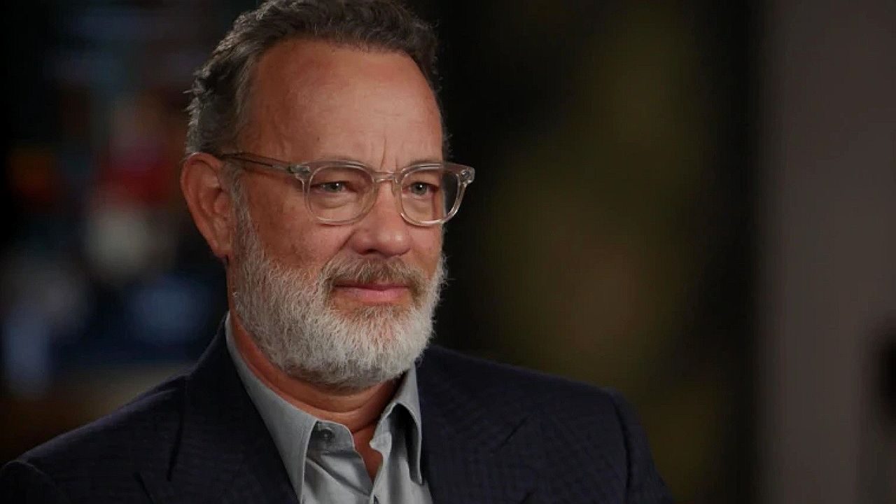 Tom Hanks durante entrevista com óculos e barba grisalha em estúdio de televisão