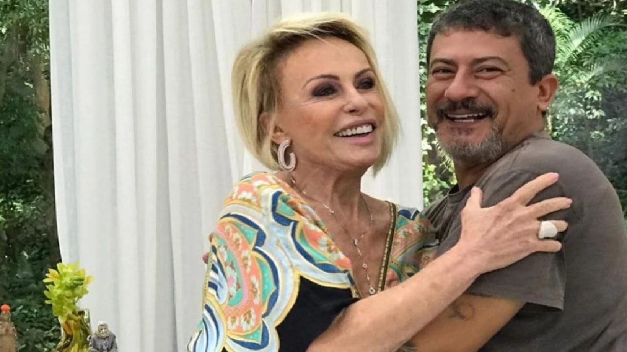 Ana Maria Braga e Tom Veiga sorrindo abraçados em ambiente com cortina branca e vegetação ao fundo