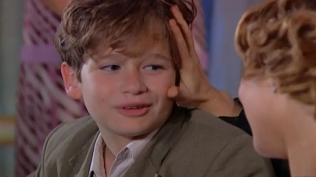 A imagem mostra um garoto de cabelos castanhos claros sendo acariciado na cabeça por uma mulher. Ele está sorrindo e usa um paletó verde-oliva com camisa clara por baixo. A cena parece ser de uma novela ou série, com um ambiente interno e cores suaves ao fundo