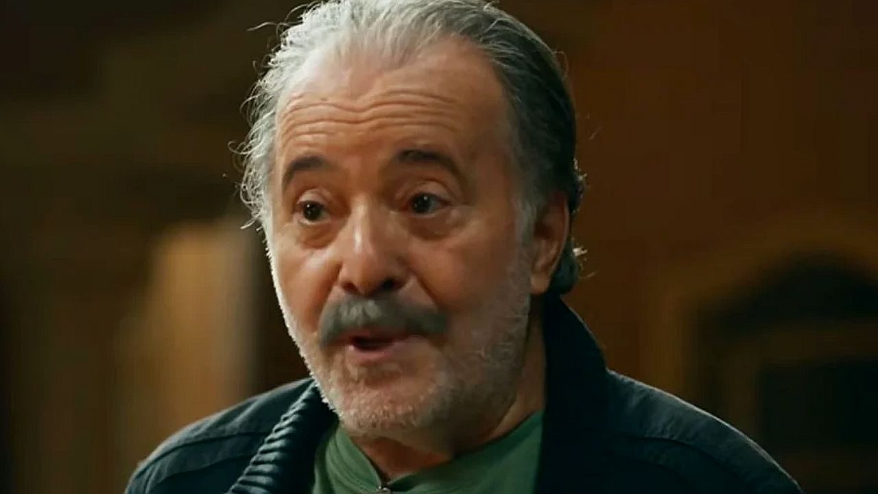 Homem grisalho de barba e bigode fala com expressão intensa durante conversa em ambiente interno iluminado em cena de novela