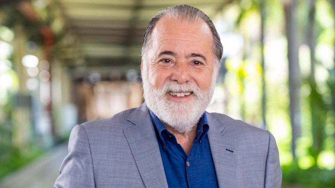 Homem idoso de barba branca e terno cinza sorri para a câmera em retrato profissional feito em corredor com área verde desfocada ao fundo