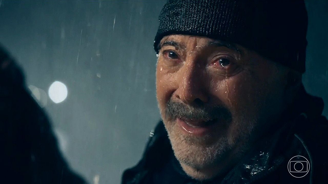 Homem usando gorro preto com rosto molhado pela chuva em cena dramática de novela da Globo