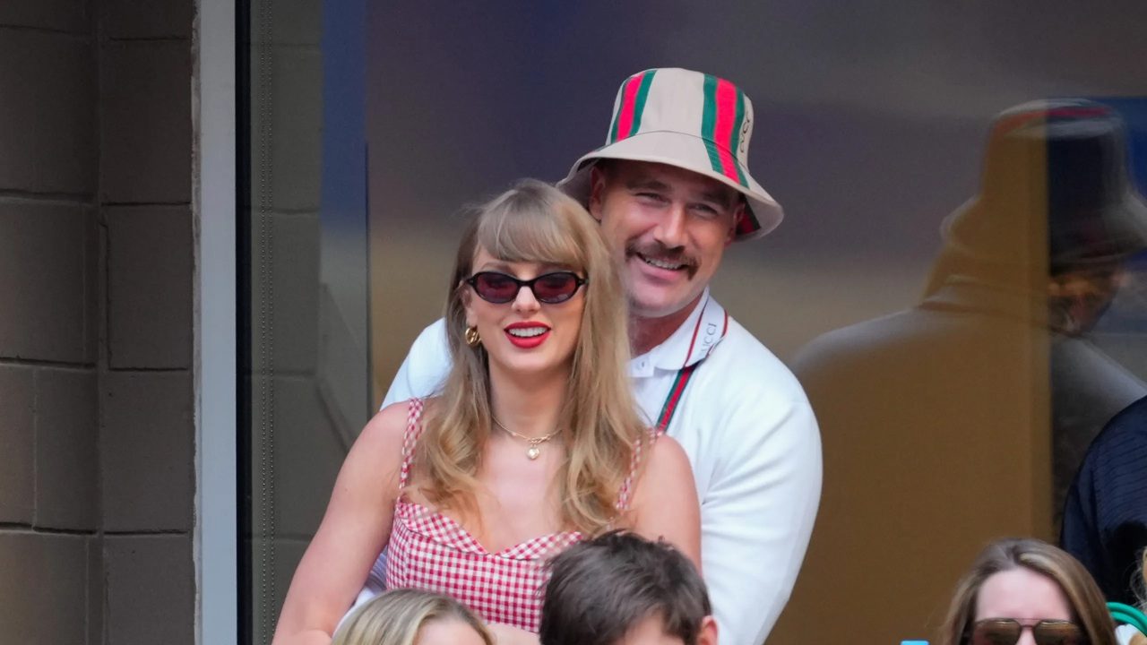 Taylor Swift e Travis Kelce sorrindo juntos em camarote durante evento esportivo