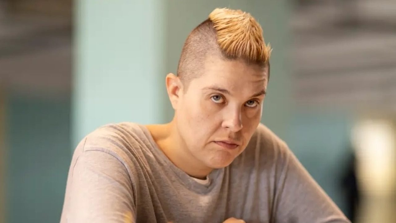 Pessoa com expressão séria olhando para a câmera, usando camiseta cinza e exibindo penteado moicano loiro em ambiente interno iluminado.