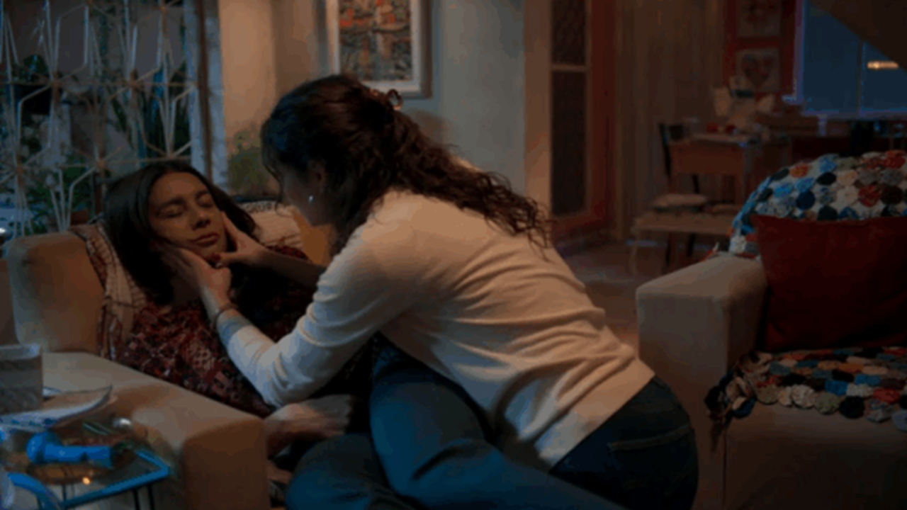 Cena mostra duas mulheres em uma sala aconchegante; uma delas está deitada no sofá com os olhos fechados, enquanto a outra, ajoelhada ao lado, segura seu rosto com preocupação, em um ambiente de iluminação suave e tom emocional