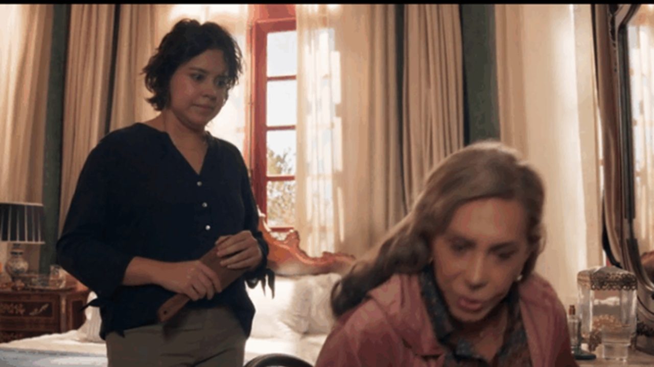 A imagem mostra duas mulheres em um quarto iluminado pela luz natural que entra por uma janela ao fundo. A mulher em pé, à esquerda, veste uma blusa escura e segura um objeto nas mãos, observando a outra, que está sentada e parece falar algo enquanto se inclina. O ambiente tem cortinas claras, uma cama e um criado-mudo decorado, transmitindo um ar doméstico e íntimo