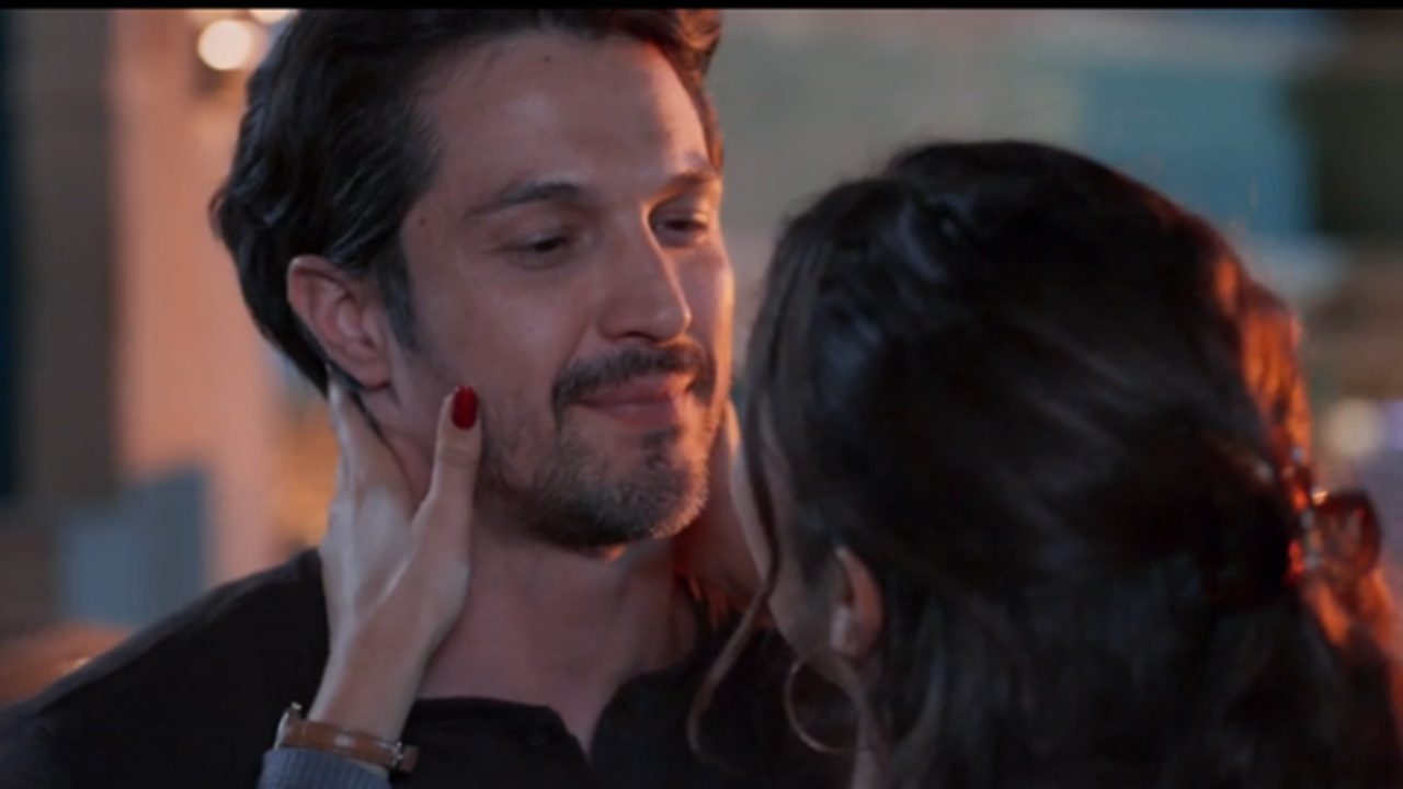 Cena mostra um homem e uma mulher em um momento íntimo e afetuoso; ela segura o rosto dele com as duas mãos, unhas pintadas de vermelho, enquanto ele sorri levemente e olha para ela com ternura em um ambiente iluminado de forma suave.