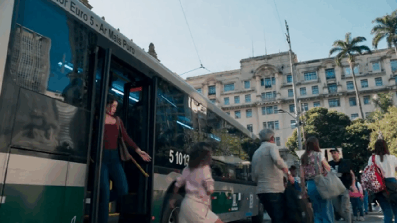 Cena da novela Três Graças mostra uma mulher descendo de um ônibus em uma movimentada área urbana, com pedestres ao redor e prédios históricos ao fundo em um dia ensolarado