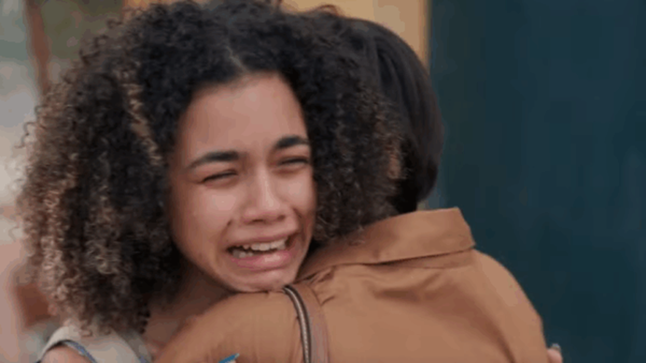 A imagem mostra uma jovem de cabelos cacheados chorando intensamente enquanto abraça outra pessoa, que veste uma camisa marrom. A expressão da jovem transmite dor e emoção, sugerindo um momento de despedida ou reencontro comovente