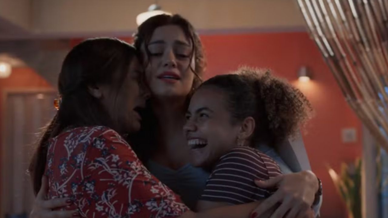 Três mulheres se abraçando em um momento de forte emoção dentro de um ambiente interno com iluminação quente, no centro uma mulher adulta com cabelos escuros presos de forma solta mantém os olhos fechados e expressão comovida enquanto envolve as outras duas com os braços, à esquerda uma mulher com cabelo longo escuro e vestido vermelho estampado apoia o rosto no ombro da mulher central demonstrando carinho e conforto, à direita uma mulher de pele mais escura com cabelo crespo preso em rabo de cavalo sorri amplamente transmitindo alívio e felicidade, o fundo mostra paredes em tom alaranjado cortinas claras e luminárias pendentes criando atmosfera acolhedora e íntima que sugere reconciliação apoio emocional ou celebração de um vínculo afetivo forte