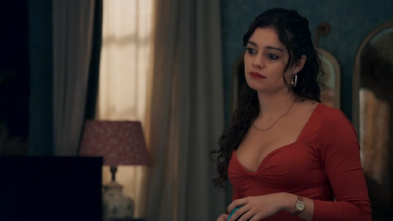 Mulher jovem de expressão séria está em um quarto com iluminação suave. Ela usa um vestido vermelho de mangas longas e decote em V, com brincos grandes e relógio no pulso. Segura um objeto pequeno nas mãos enquanto observa algo fora de quadro. Ao fundo, cortinas claras filtram a luz da janela, e uma luminária com cúpula rosada ilumina parte do ambiente