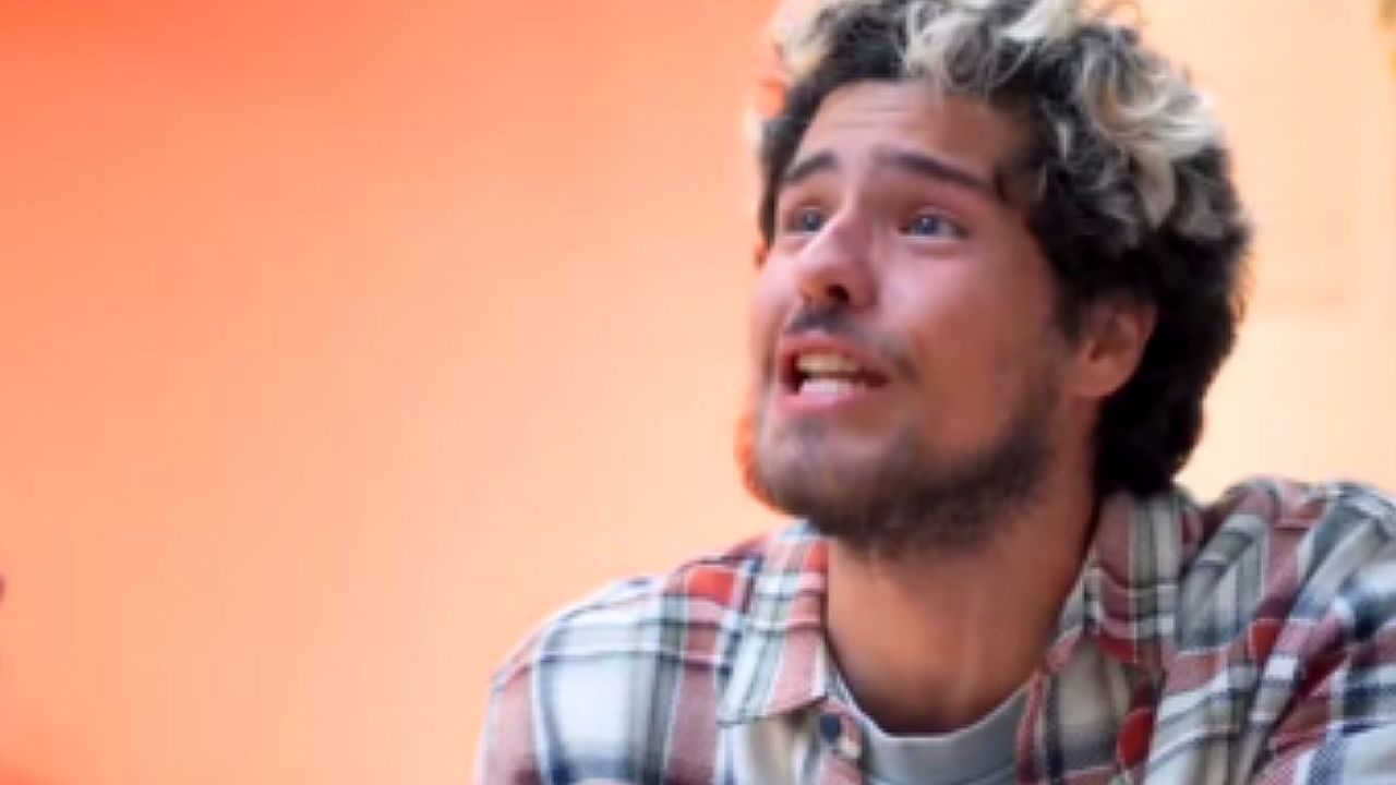 Homem jovem de cabelos cacheados com mechas claras e barba rala fala com expressão exaltada, olhando para cima durante conversa em ambiente externo com fundo alaranjado desfocado