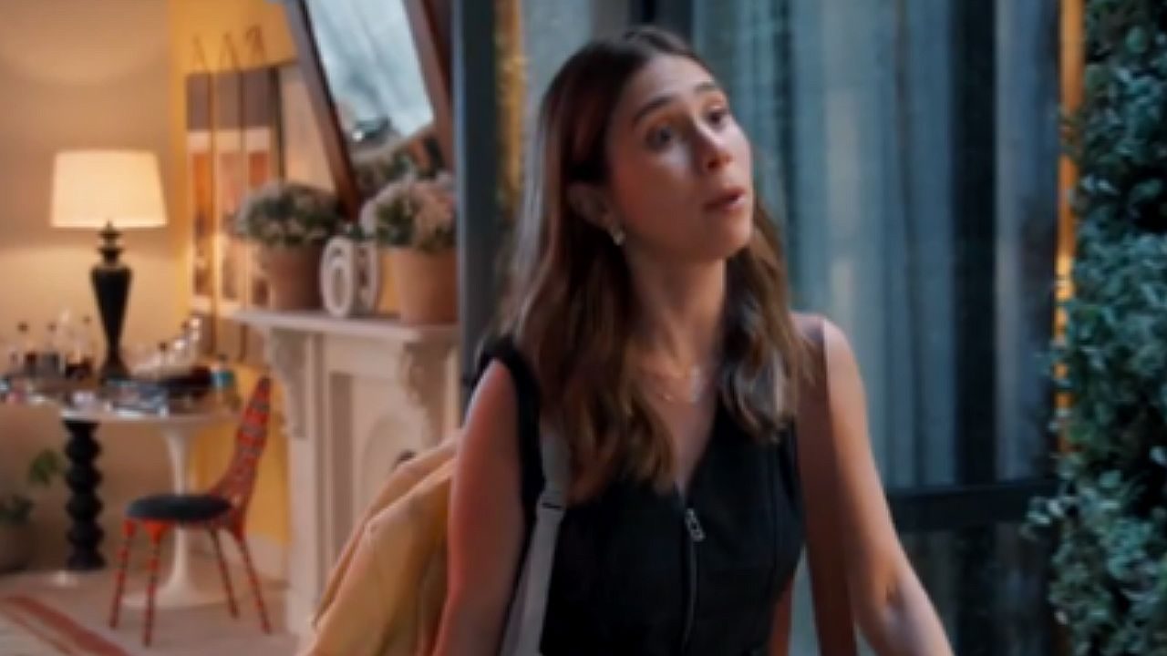 Mulher com mochila fala na porta de casa em cena de novela com sala iluminada ao fundo