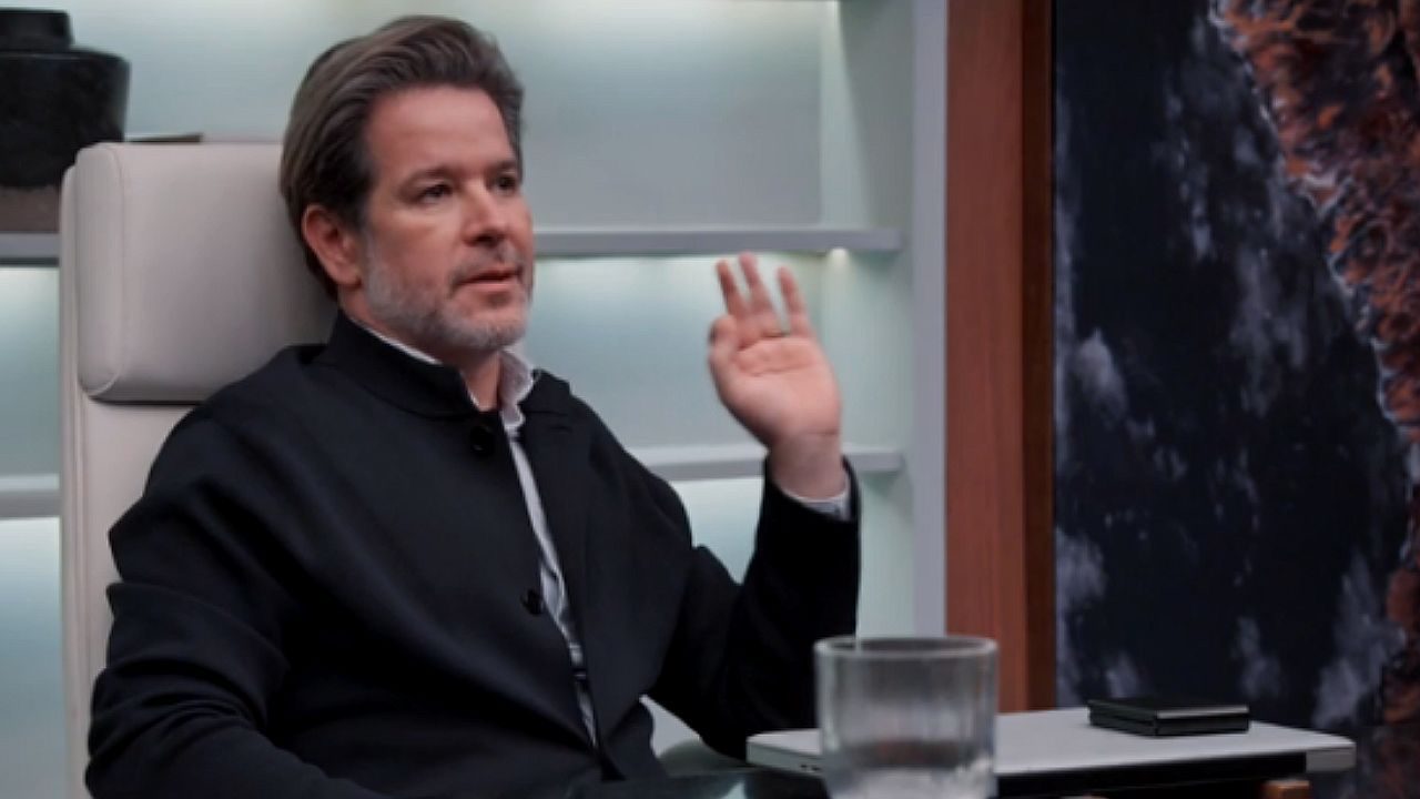 Homem de terno gesticula enquanto conversa sentado em escritório moderno com tablet sobre a mesa