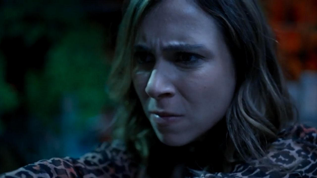 Mulher com expressão tensa encara algo fora de quadro em cena noturna de suspense com iluminação azul e verde