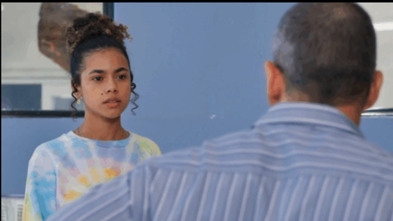 Jovem de cabelo cacheado preso em um coque alto olha séria para um homem de costas. Ela veste uma blusa colorida em tie-dye, enquanto o homem usa uma camisa listrada azul. Ambos estão em um ambiente interno de paredes azuis.