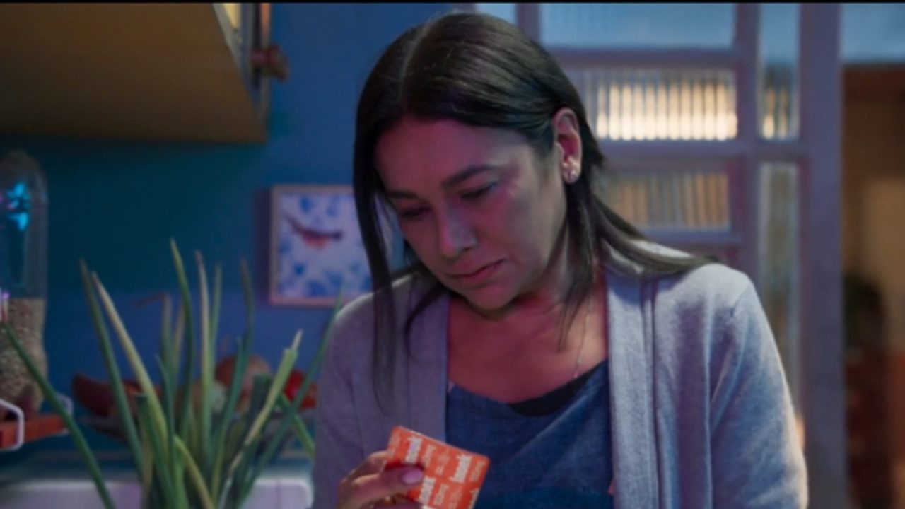 Mulher em ambiente doméstico pouco iluminado olha para uma caixa de medicamento laranja com expressão triste e cansada. Ela veste cardigã cinza sobre blusa azul, e ao fundo há plantas e objetos de cozinha desfocados, compondo um clima de preocupação ou reflexão.