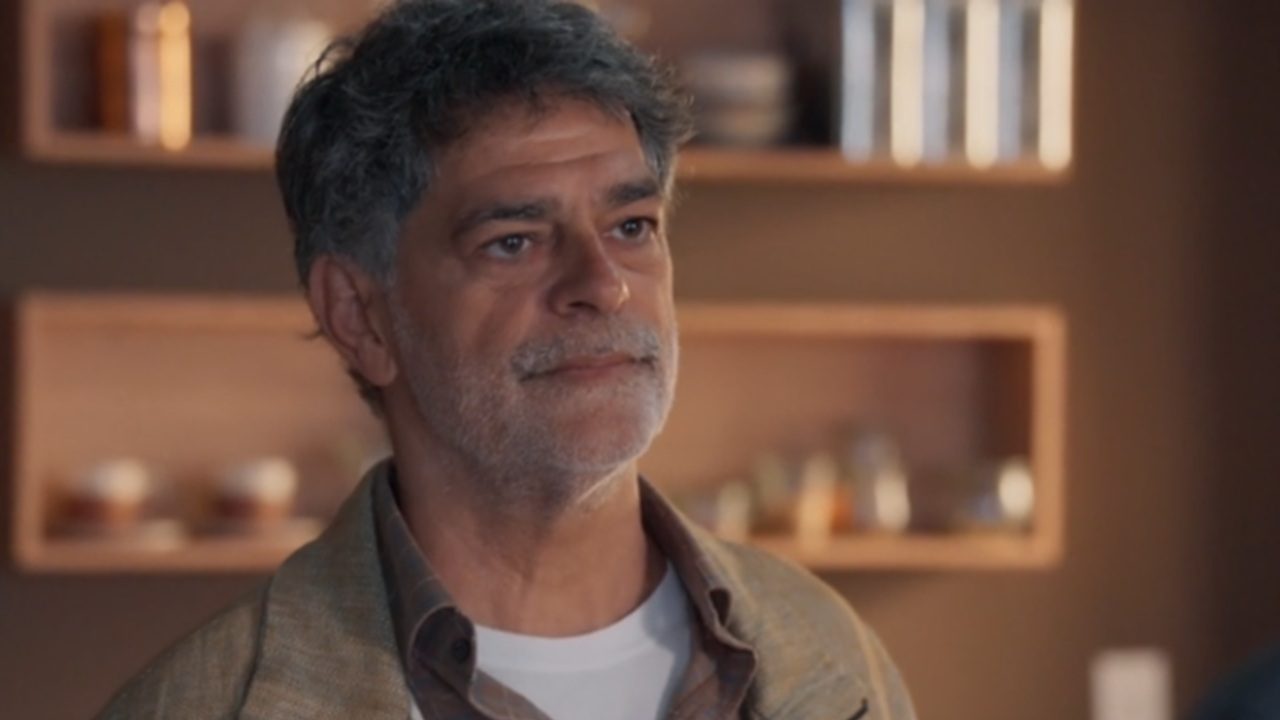 Homem de meia-idade aparece em um ambiente interno com iluminação quente e aconchegante. Ele tem cabelos grisalhos, barba por fazer e expressão serena, olhando levemente para a frente. Usa uma camiseta branca por baixo de uma camisa marrom e um casaco em tom neutro. Ao fundo, há prateleiras embutidas com objetos decorativos e iluminação suave, criando um clima acolhedor e sofisticado.